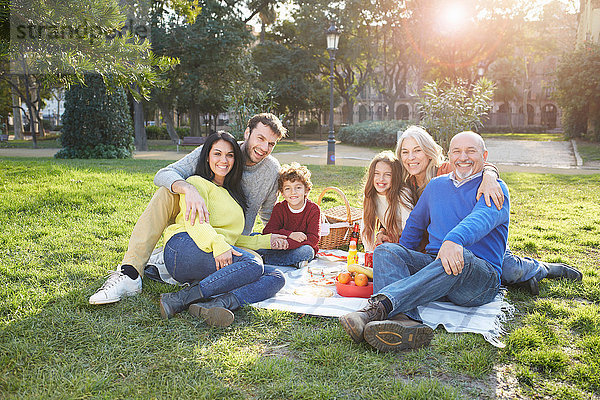 Mehrgenerationen-Familie sitzt auf dem Rasen beim Picknick und schaut lächelnd in die Kamera