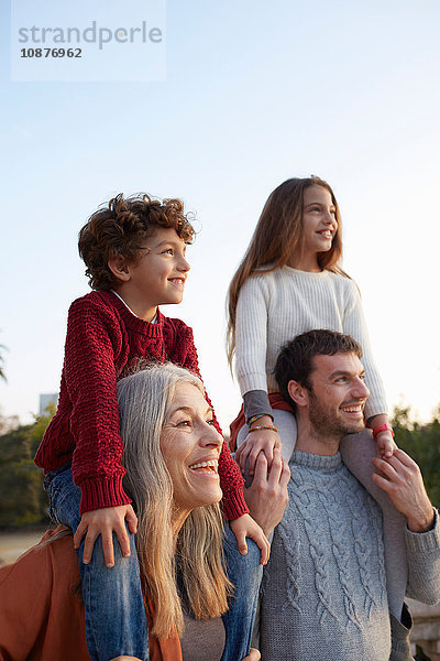 Mädchen und Junge sitzen auf den Schultern von Großmutter und Vater und schauen lächelnd weg