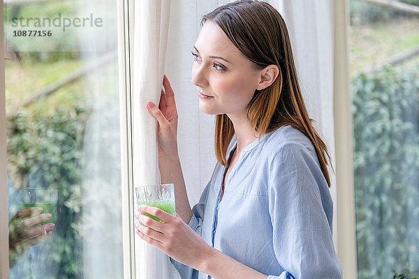 Frau schaut aus dem Fenster und hält grünen Smoothie
