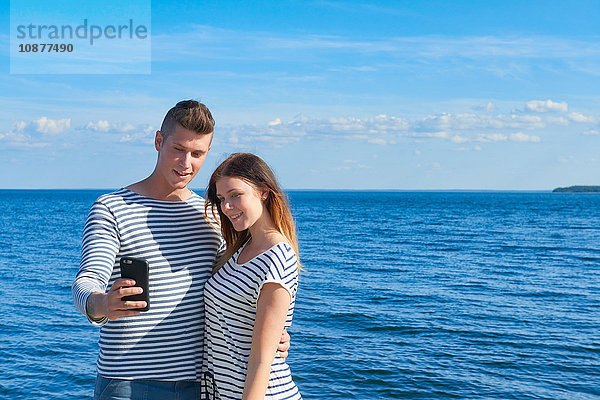 Junges Paar steht am Meer und fotografiert sich selbst mit einem Smartphone