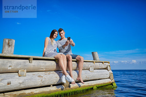 Ehepaar sitzt auf einem Holzsteg und macht ein Selbstporträt mit einem Smartphone