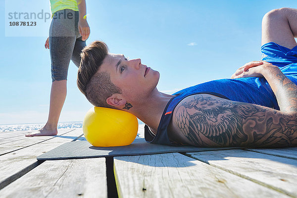 Tätowierter Mann liegt auf Pier und benutzt Ball als Kissen