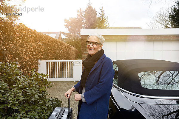 Frau in der Nähe des Autos auf der Einfahrt mit einem Koffer in der Hand und lächelndem Blick in die Kamera