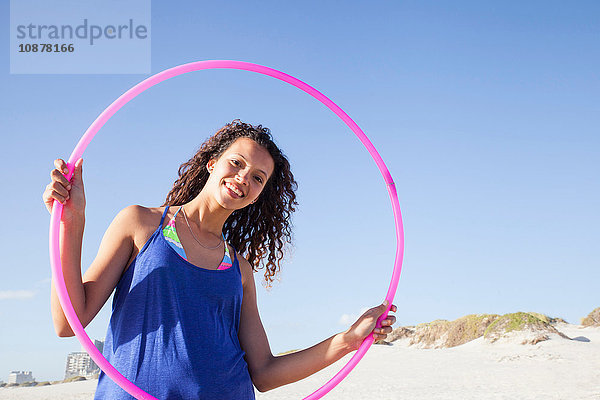 Frau am Strand schaut durch einen Hula-Hoop-Reifen in die Kamera und lächelt