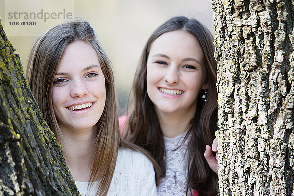 Porträt von Freundinnen hinter Baumstämmen  die lächelnd in die Kamera schauen