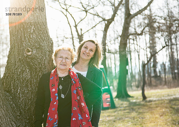 Erwachsene Tochter steht hinter der Mutter am Baum und schaut lächelnd in die Kamera