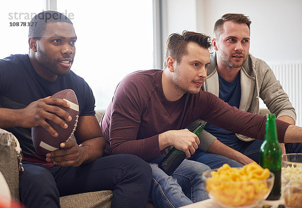 Gruppe von Männern  die einen Ball halten und ein Sportereignis im Fernsehen mit Snacks und Bieren verfolgen