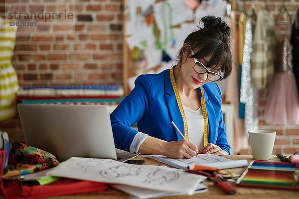 Frau im Design-Studio sitzt am Schreibtisch und skizziert Modedesign