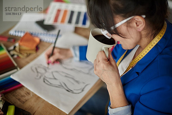 Über-Schulter-Ansicht einer Frau im Designstudio  die Kaffee trinkt und Modedesign skizziert