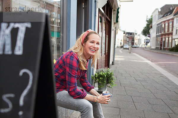 Frau sitzt auf der Schaufensterbank  hält Kaffeetasse und schaut lächelnd weg