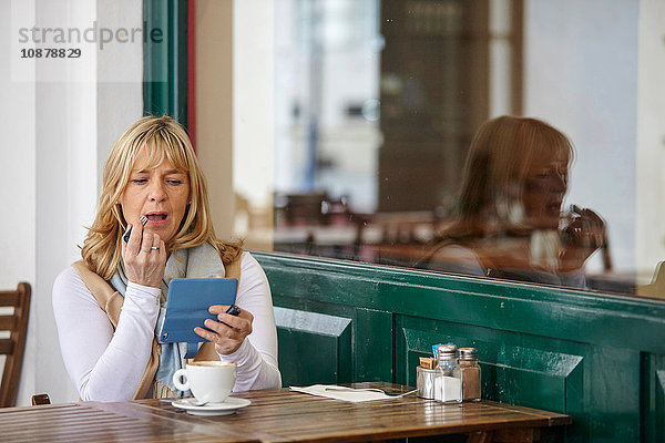 Reife Frau benutzt Smartphone zum Auftragen von Lippenstift am Tisch im Café auf dem Bürgersteig