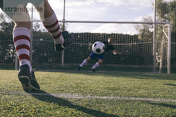 Legs of a footnball player kicking a ball in front of a goal with a goalkeeper