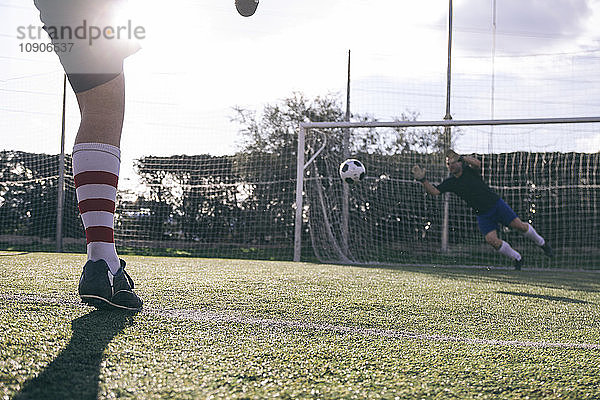 Legs of a footnball player kicking a ball in front of a goal with a goalkeeper