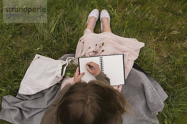 Junge Frau sitzt auf einer Wiese und schreibt etwas in ihr Notizbuch.