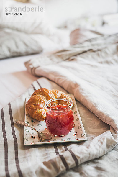 Teller mit Croissant und Glas Erdbeermarmelade auf dem Bett