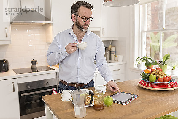 Man with cup of coffee standing in the kitchen using digital tablet