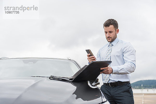 Businessman with documents and cell phone at car