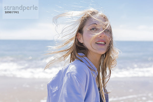 Porträt einer lächelnden jungen Frau mit blasenden Haaren am Meer