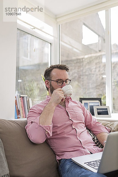 Mature man on couch drinking coffee and using laptop
