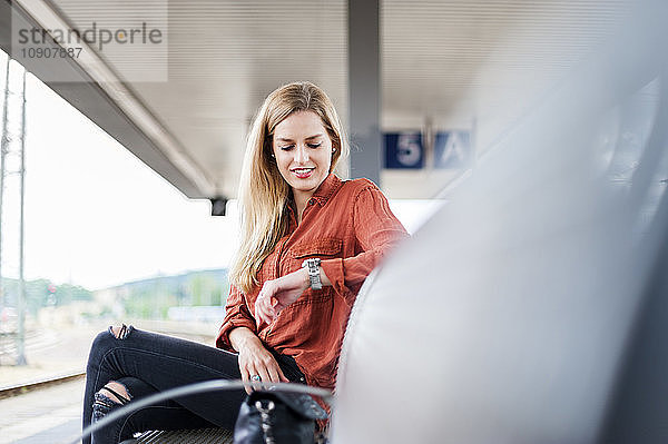 Smiling young woman sitting on bench at platform checking the time