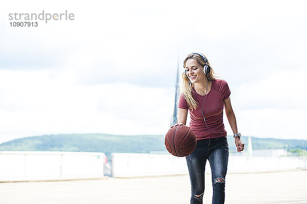 Smiling young woman with headphones playing with basketball on roof terrace