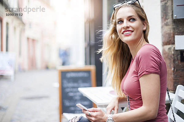 Porträt einer lächelnden Frau mit einem Smartphone  das in einem Straßencafé sitzt und etwas beobachtet.