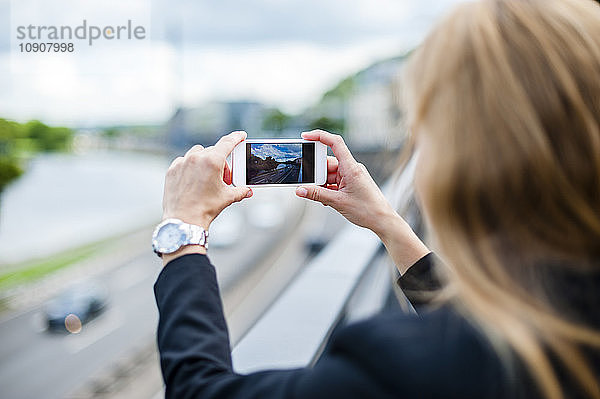 Back view of woman standing on a bridge taking photo with smartphone
