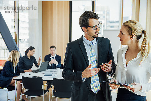 Businessman and businesswoman talking in office with meeting in background