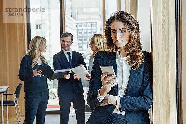 Businesswoman in office looking at cell phone with businesspeople in background
