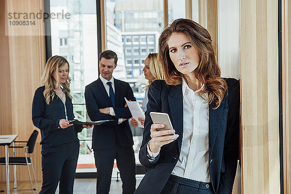 Businesswoman in office holding cell phone with businesspeople in background
