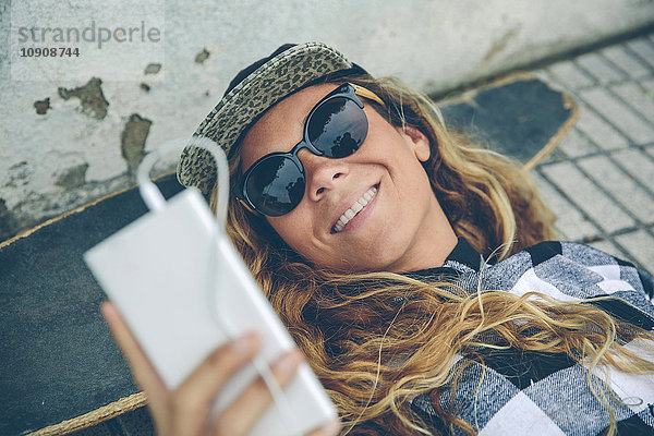 Lächelnde junge Frau liegt auf dem Skateboard und schaut aufs Handy.