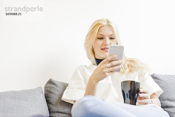 Blonde junge Frau sitzt auf der Couch mit einer Tasse Kaffee und schaut auf das Smartphone.