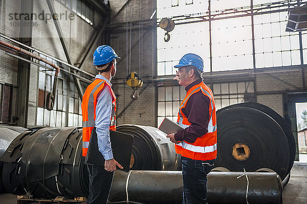 Two men with safety vests in factory hall with rolls of rubber