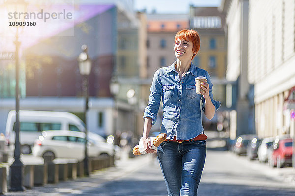 Lächelnde junge Frau  die mit Kaffee und Baguette in der Stadt spazieren geht.