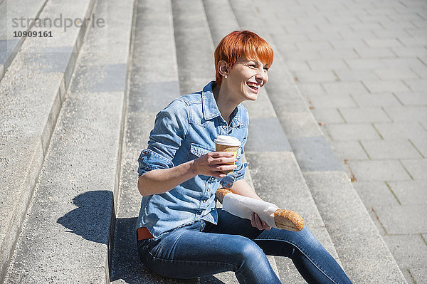 Fröhliche junge Frau sitzt auf der Treppe mit Kaffee zum Mitnehmen