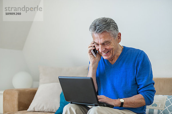 Lächelnder älterer Mann sitzt auf der Couch mit Laptop und Handy