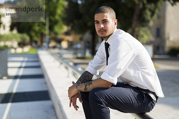 Porträt eines jungen Geschäftsmannes mit Tätowierungen auf den Unterarmen auf dem Geländer sitzend