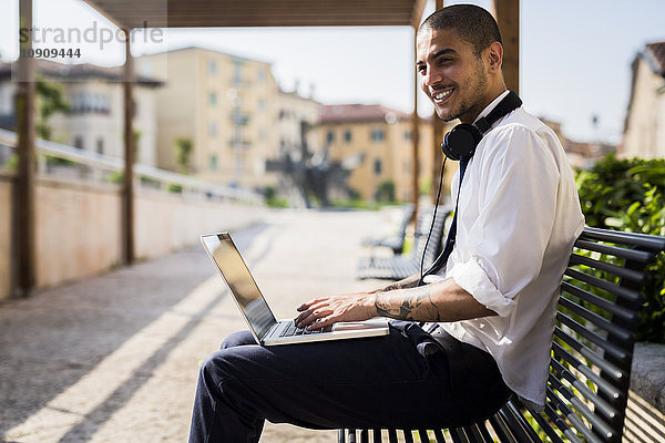 Lächelnder junger Geschäftsmann auf einer Bank sitzend mit Laptop