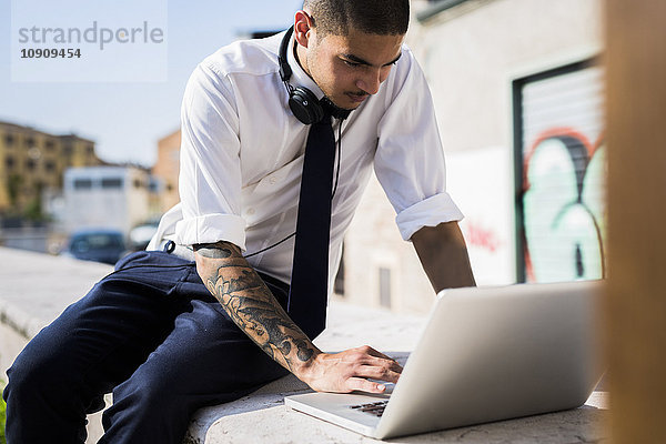 Junger Geschäftsmann sitzt auf einer Wand mit Laptop