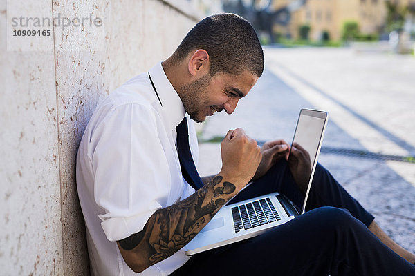 Glücklicher junger Geschäftsmann sitzt auf dem Boden und schaut auf seinen Laptop.