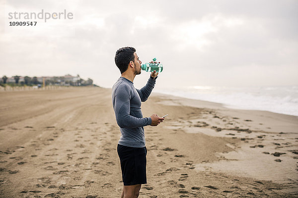 Sportlicher junger Mann beim Trinken aus der Flasche am Strand