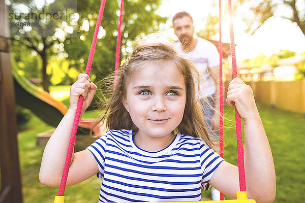 Porträt eines glücklichen kleinen Mädchens auf einer Schaukel mit Vater im Hintergrund