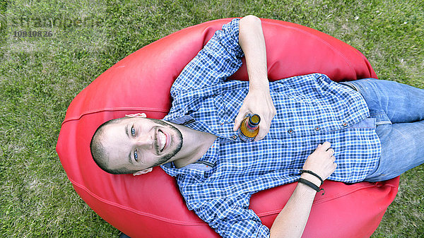 Mann entspannt auf Bohnensack im Garten beim Biertrinken