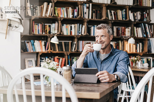 Erwachsener Mann sitzt im Café mit digitaler Tablette und trinkt Kaffee.