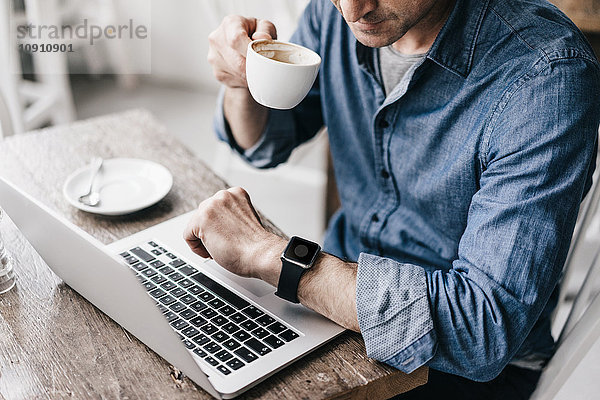 Reifer Mann  der Kaffee trinkt  während er am Laptop arbeitet  mit Blick auf smartwatch