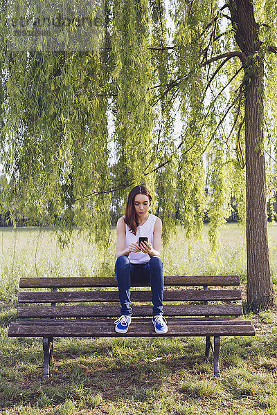 Junge Frau mit Telefon sitzt auf einer Bank im Park