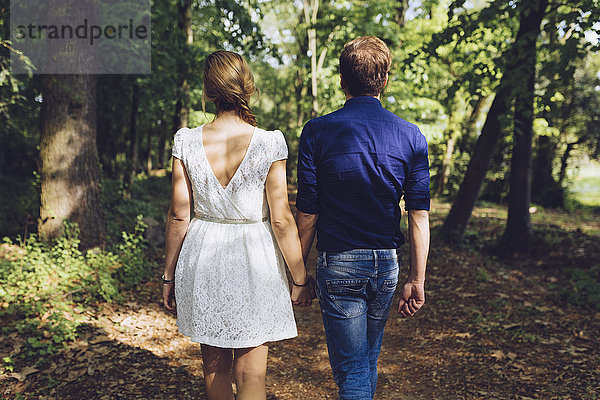 Rückansicht eines verliebten Paares  das Hand in Hand im Wald spazieren geht.
