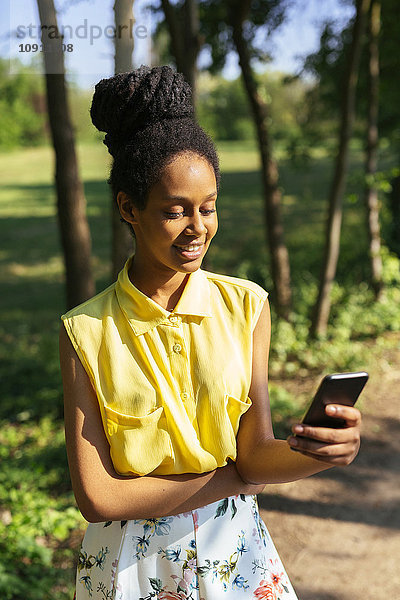 Porträt einer lächelnden jungen Frau in der Natur mit Blick auf ihr Smartphone