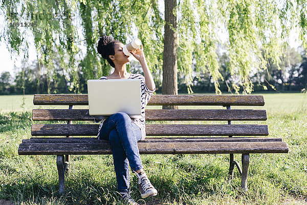 Junge Frau mit Laptop auf Parkbank sitzend  Kaffee trinkend zum Mitnehmen