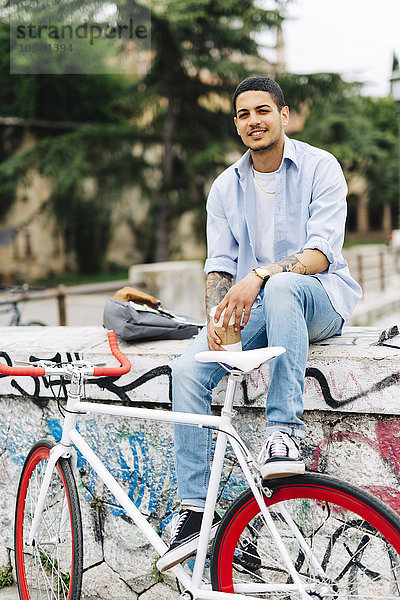 Lächelnder junger Mann mit Fahrrad auf Graffiti-Wand sitzend